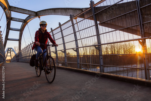 Caucasian Woman Riding a Bicycle on a Pedestrian Bridge over the Highway during a sunny sunset. Taken in Surrey, Vancouver, British Columbia, Canada.