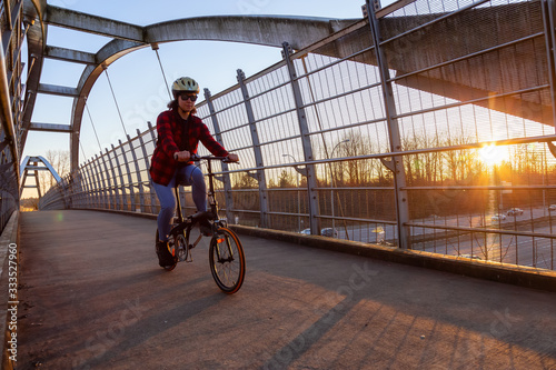 Fototapeta Naklejka Na Ścianę i Meble -  Caucasian Woman Riding a Bicycle on a Pedestrian Bridge over the Highway during a sunny sunset. Taken in Surrey, Vancouver, British Columbia, Canada.