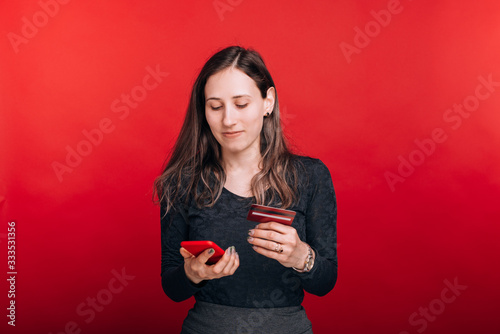 Beautiful young woman si looking at her phone, buying something online and paying with a credit card on red background.