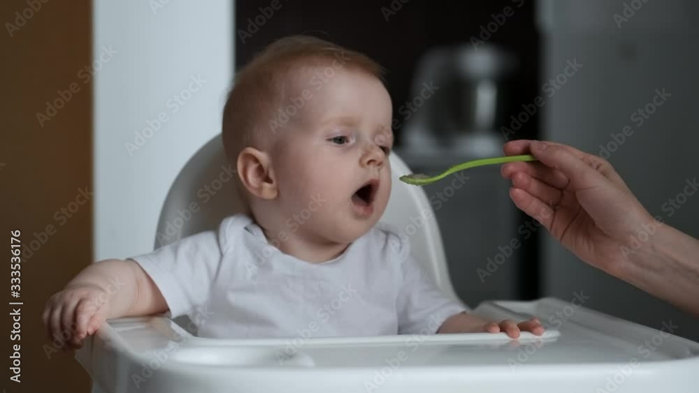 Cute little baby feeding with a spoon at the table, the baby smiles at the camera and then cries and gets cranky infant eating meal foods. 4k 50 fps Cute little baby feeding with a spoon at the table, the baby smiles at the camera and then cries and gets cranky infant eating meal foods. 4k 50 fps