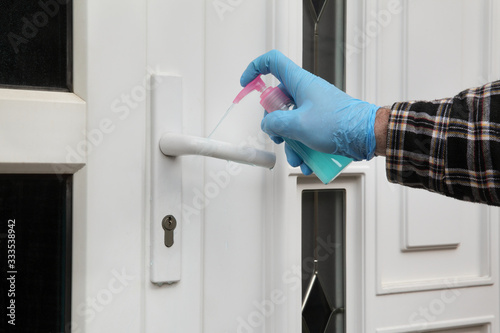 Huna hand in protective glove spraying antibacterial or cleaning solution from sprayer bottle to handle of house door photo