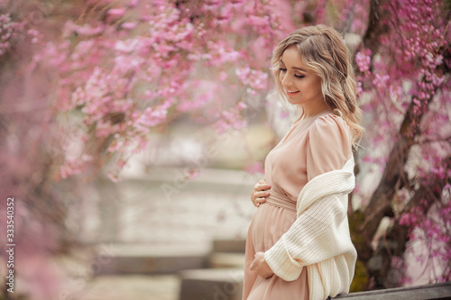 Young beautiful blonde pregnant girl in a pink dress on a walk in the park under a sakura tree.