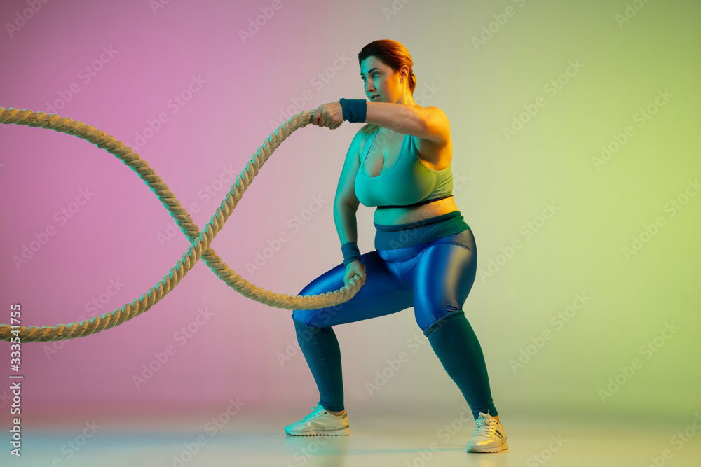Obraz premium Young caucasian plus size female model's training on gradient purple green background in neon light. Doing workout exercises with ropes. Concept of sport, healthy lifestyle, body positive, equality.