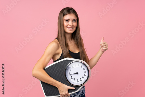 Young girl with weighing machine and with thumb up over isolated pink background