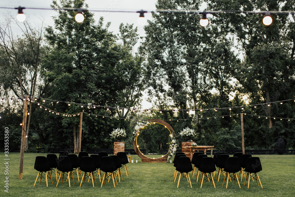 Wooden wedding arch decorated by white flowers with greenery standing in the center of wedding ceremony. Black chairs on sides. Cozy rustic wedding ceremony with flower arch in garden beautiful.