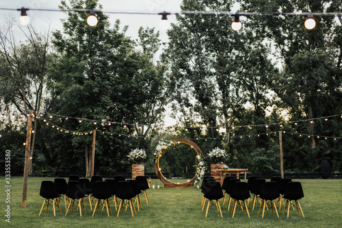 Obraz na płótnie Wooden wedding arch decorated by white flowers with greenery standing in the center of wedding ceremony. Black chairs on sides. Cozy rustic wedding ceremony with flower arch in garden beautiful. w salonie