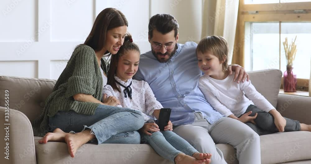 Little schoolgirl relaxing on sofa with smiling parents and younger brother, showing funny video on smartphone. Full length happy family using mobile applications, enjoying leisure time together. Little schoolgirl relaxing on sofa with smiling parents and younger brother, showing funny video on smartphone. Full length happy family using mobile applications, enjoying leisure time together.