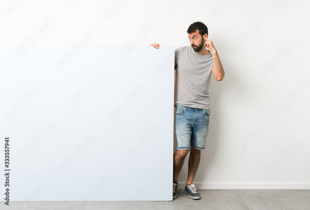 Young handsome man with beard holding a big blue empty placard having doubts and thinking Young handsome man with beard holding a big blue empty placard having doubts and thinking