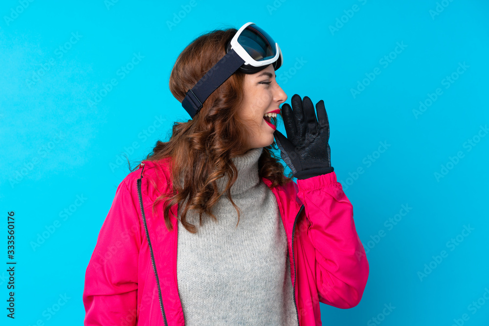Skier woman with snowboarding glasses over isolated blue wall shouting with mouth wide open