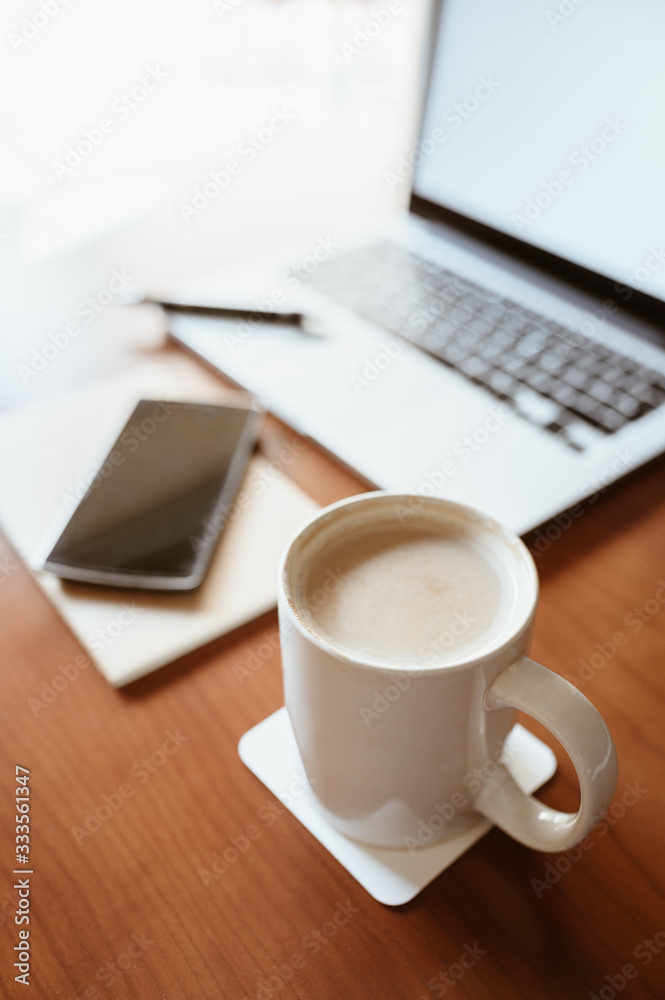 White cup of coffee with laptop and smart phone on wooden office desk, midday light comes in through the window. Business and home concept White cup of coffee with laptop and smart phone on wooden office desk, midday light comes in through the window. Business and home concept