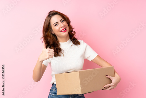 Young Russian woman over isolated pink background holding a box to move it to another site with thumb up © luismolinero