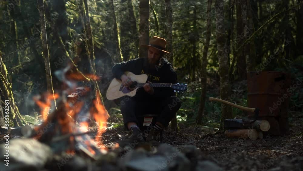 Young man in hat with guitar playing songs and singing next to the campfire in dense rainforest Young man in hat with guitar playing songs and singing next to the campfire in dense rainforest