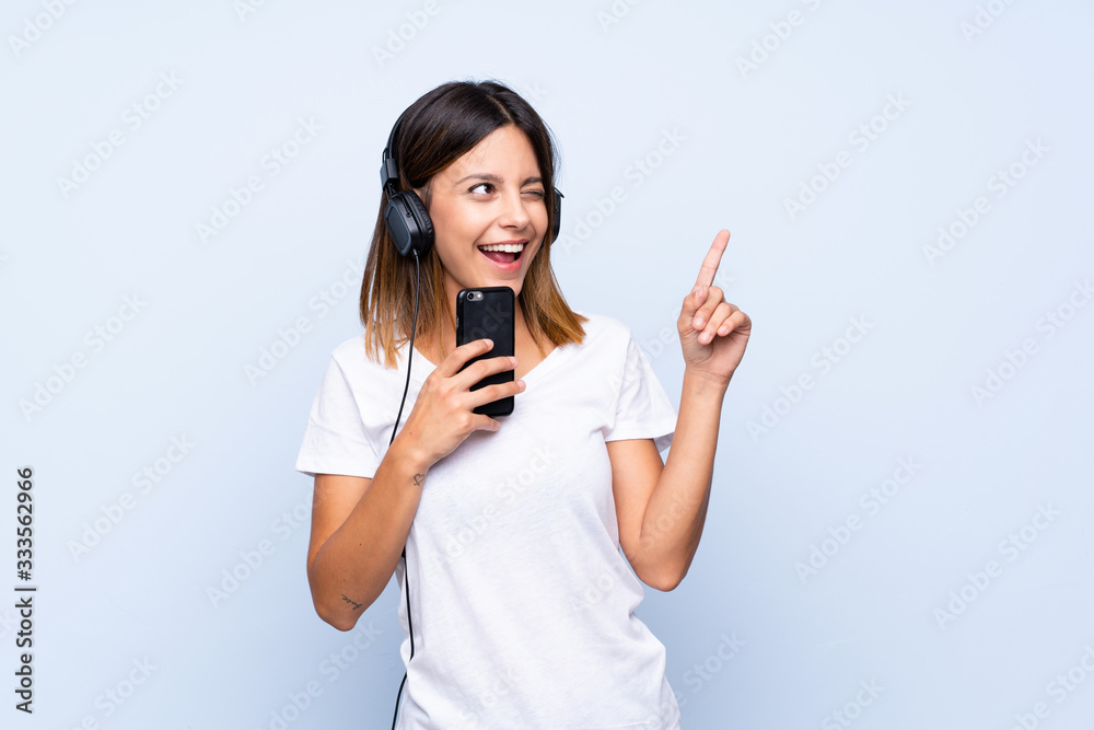 Young woman over isolated blue background using the mobile with headphones and singing Young woman over isolated blue background using the mobile with headphones and singing