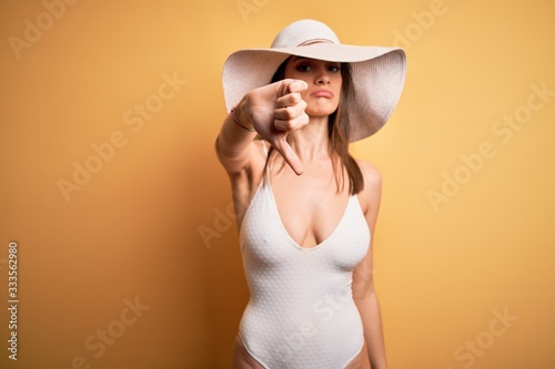 Young beautiful brunette woman on vacation wearing swimsuit and summer hat looking unhappy and angry showing rejection and negative with thumbs down gesture. Bad expression.