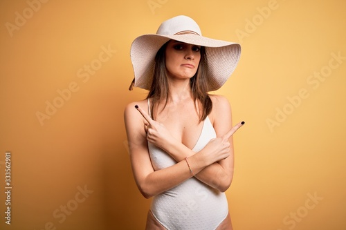 Young beautiful brunette woman on vacation wearing swimsuit and summer hat Pointing to both sides with fingers, different direction disagree