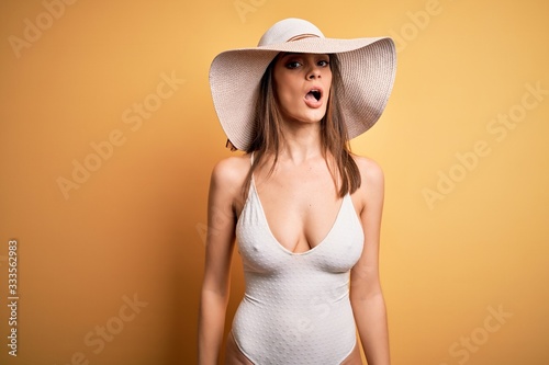 Young beautiful brunette woman on vacation wearing swimsuit and summer hat In shock face, looking skeptical and sarcastic, surprised with open mouth