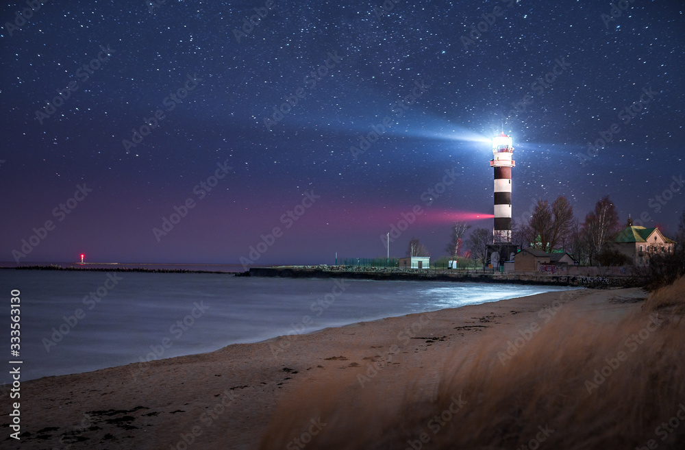 Majestyc lighthouse with beacon of light next to sea at night time full of stars. 