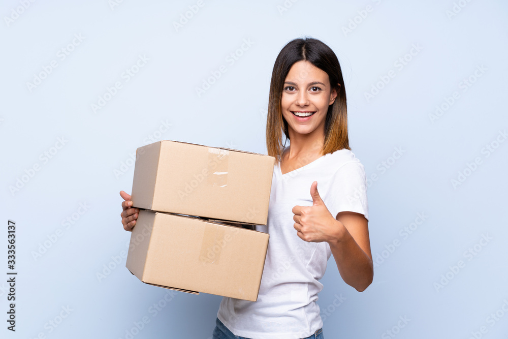 Young woman over isolated blue background holding a box to move it to another site with thumb up Young woman over isolated blue background holding a box to move it to another site with thumb up