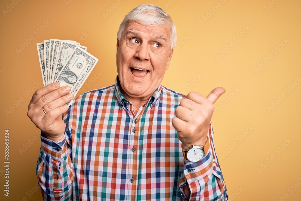 Senior handsome hoary man holding bunch of dollars banknotes over yellow background pointing and showing with thumb up to the side with happy face smiling Senior handsome hoary man holding bunch of dollars banknotes over yellow background pointing and showing with thumb up to the side with happy face smiling
