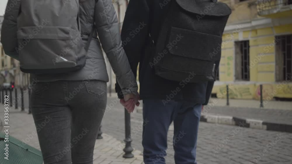 Young Caucasian man and woman taking each other's hands and walking away along town street. Boyfriend and girlfriend with backpacks dating in city. Adolescence, romance, love, lifestyle. Young Caucasian man and woman taking each other's hands and walking away along town street. Boyfriend and girlfriend with backpacks dating in city. Adolescence, romance, love, lifestyle.