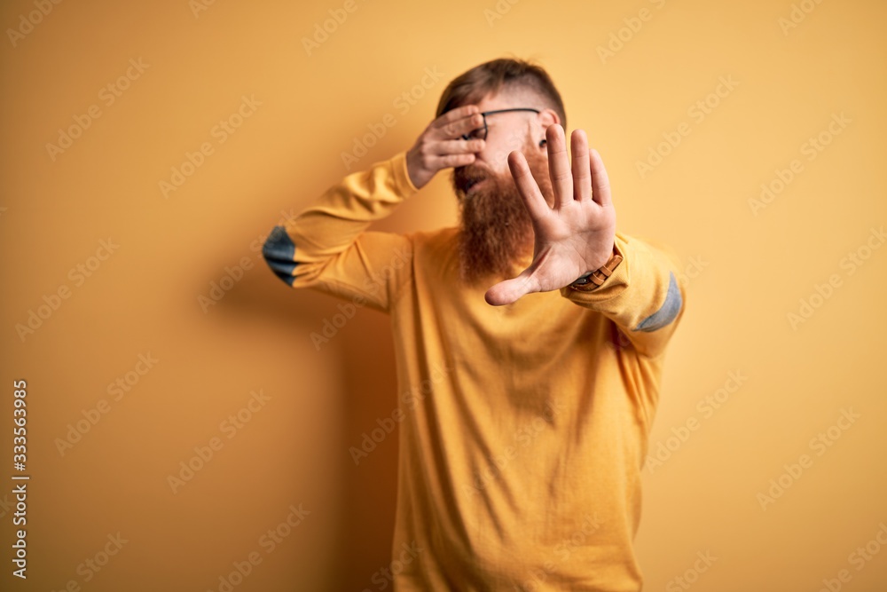 Handsome Irish redhead man with beard wearing glasses over yellow isolated background covering eyes with hands and doing stop gesture with sad and fear expression. Embarrassed and negative concept. Handsome Irish redhead man with beard wearing glasses over yellow isolated background covering eyes with hands and doing stop gesture with sad and fear expression. Embarrassed and negative concept.