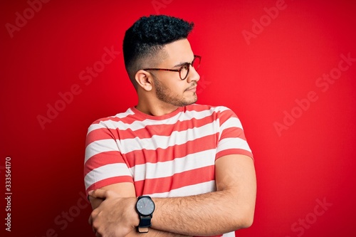 Young handsome man wearing casual striped t-shirt and glasses over isolated red background looking to the side with arms crossed convinced and confident