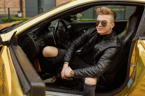 A businessman is sitting in a golden car. A man is driving inside a car. Driver. Chic man with glasses, with a perfect haircut, glasses, fashionable. Freelancer. Rich boy. A teenager drives a car.