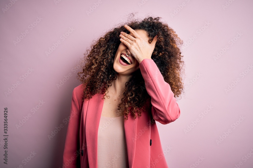 Young beautiful businesswoman with curly hair and piercing wearing elegant jacket smiling and laughing with hand on face covering eyes for surprise. Blind concept.