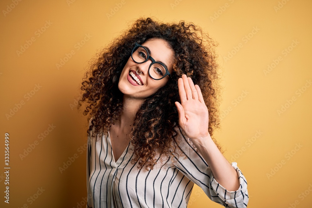 Young beautiful woman with curly hair and piercing wearing striped shirt and glasses Waiving saying hello happy and smiling, friendly welcome gesture Young beautiful woman with curly hair and piercing wearing striped shirt and glasses Waiving saying hello happy and smiling, friendly welcome gesture