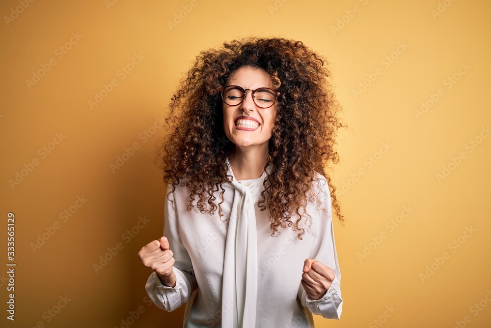 Young beautiful brunette woman with curly hair and piercing wearing shirt and glasses very happy and excited doing winner gesture with arms raised, smiling and screaming for success. Celebration Young beautiful brunette woman with curly hair and piercing wearing shirt and glasses very happy and excited doing winner gesture with arms raised, smiling and screaming for success. Celebration