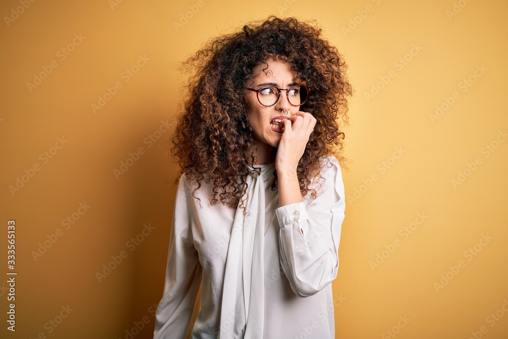 Young beautiful brunette woman with curly hair and piercing wearing shirt and glasses looking stressed and nervous with hands on mouth biting nails. Anxiety problem. Young beautiful brunette woman with curly hair and piercing wearing shirt and glasses looking stressed and nervous with hands on mouth biting nails. Anxiety problem.