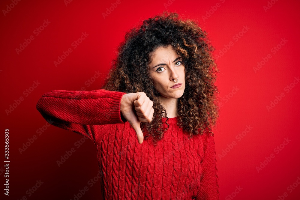 Young beautiful woman with curly hair and piercing wearing casual red sweater looking unhappy and angry showing rejection and negative with thumbs down gesture. Bad expression. Young beautiful woman with curly hair and piercing wearing casual red sweater looking unhappy and angry showing rejection and negative with thumbs down gesture. Bad expression.