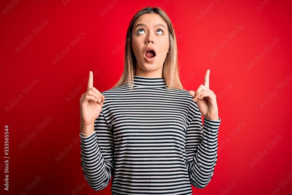 Young beautiful blonde woman wearing casual striped sweater over red isolated background amazed and surprised looking up and pointing with fingers and raised arms. Young beautiful blonde woman wearing casual striped sweater over red isolated background amazed and surprised looking up and pointing with fingers and raised arms.