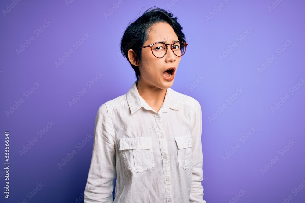 Young beautiful asian girl wearing casual shirt and glasses standing over purple background In shock face, looking skeptical and sarcastic, surprised with open mouth Young beautiful asian girl wearing casual shirt and glasses standing over purple background In shock face, looking skeptical and sarcastic, surprised with open mouth