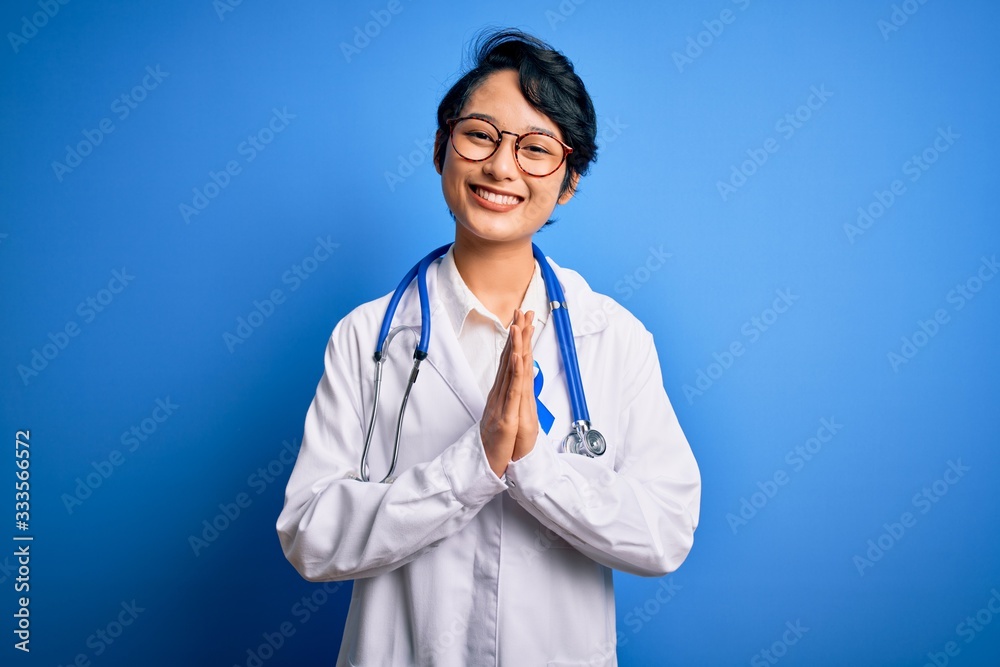 Young beautiful asian doctor girl wearing stethoscope and coat with blue cancer ribbon praying with hands together asking for forgiveness smiling confident. Young beautiful asian doctor girl wearing stethoscope and coat with blue cancer ribbon praying with hands together asking for forgiveness smiling confident.