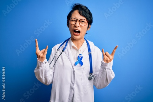 Young beautiful asian doctor girl wearing stethoscope and coat with blue cancer ribbon shouting with crazy expression doing rock symbol with hands up. Music star. Heavy concept.