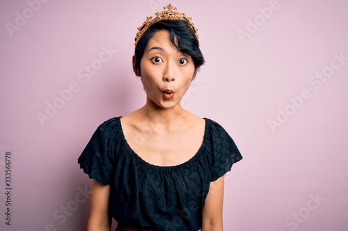 Fotografie Young beautiful chinese woman wearing golden crown of king over isolated pink background afraid and shocked with surprise expression, fear and excited face
