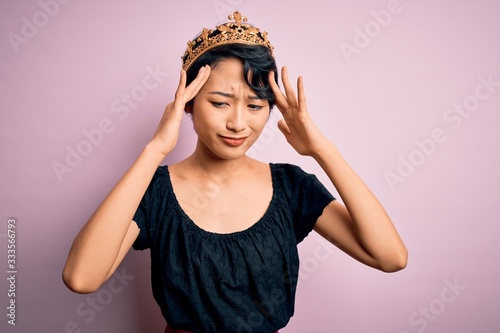 Young beautiful chinese woman wearing golden crown of king over isolated pink background suffering from headache desperate and stressed because pain and migraine. Hands on head.