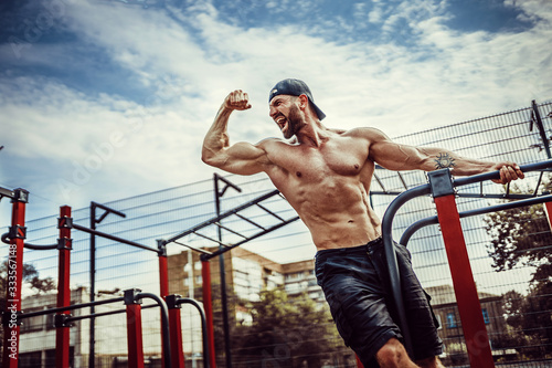 Bearded bodybuilder man exercising on monkey bars for the upper-body in a modern calisthenics park outdoors on a sunny day. Show biceps and scream for motivation.