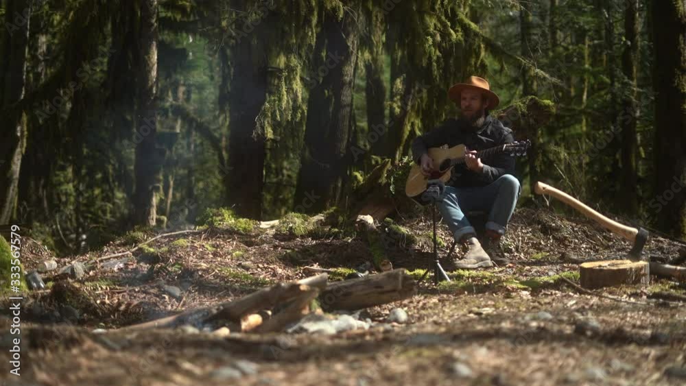 Young man in hat with guitar playing songs and singing next to the campfire in dense rainforest Young man in hat with guitar playing songs and singing next to the campfire in dense rainforest