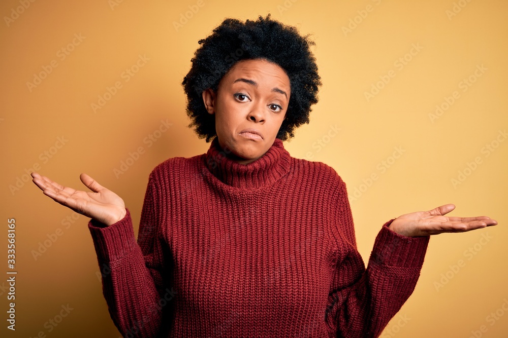 Young beautiful African American afro woman with curly hair wearing casual turtleneck sweater clueless and confused expression with arms and hands raised. Doubt concept. Young beautiful African American afro woman with curly hair wearing casual turtleneck sweater clueless and confused expression with arms and hands raised. Doubt concept.
