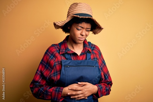 Young African American afro farmer woman with curly hair wearing apron and hat with hand on stomach because indigestion, painful illness feeling unwell. Ache concept.