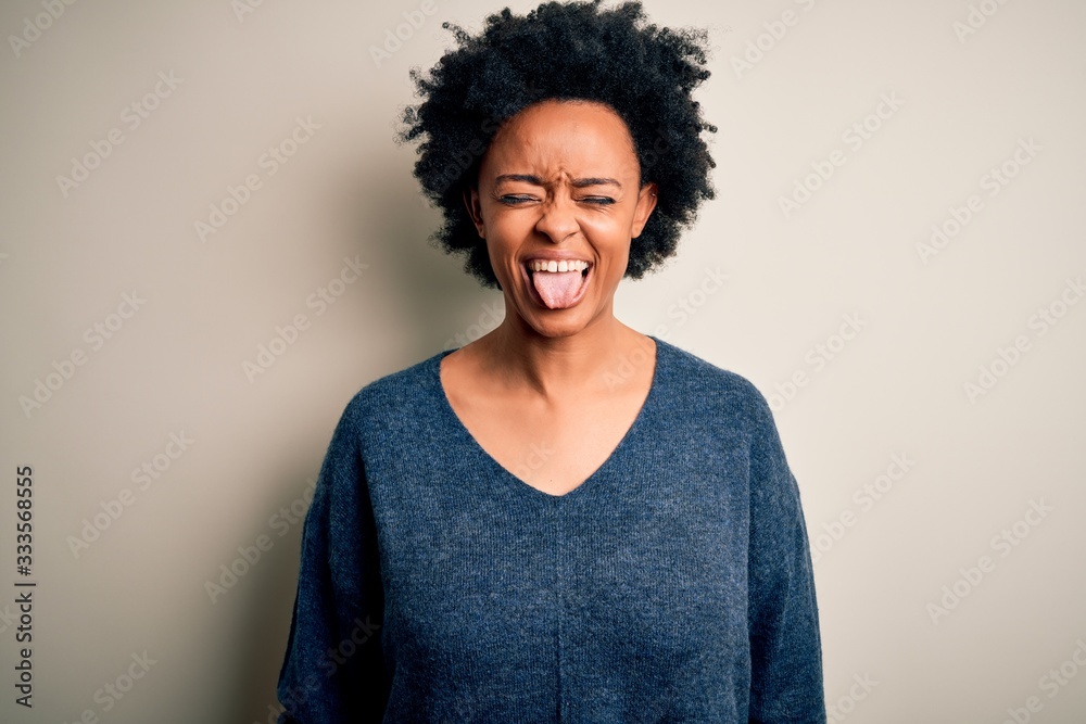 Young beautiful African American afro woman with curly hair wearing casual sweater sticking tongue out happy with funny expression. Emotion concept. Young beautiful African American afro woman with curly hair wearing casual sweater sticking tongue out happy with funny expression. Emotion concept.