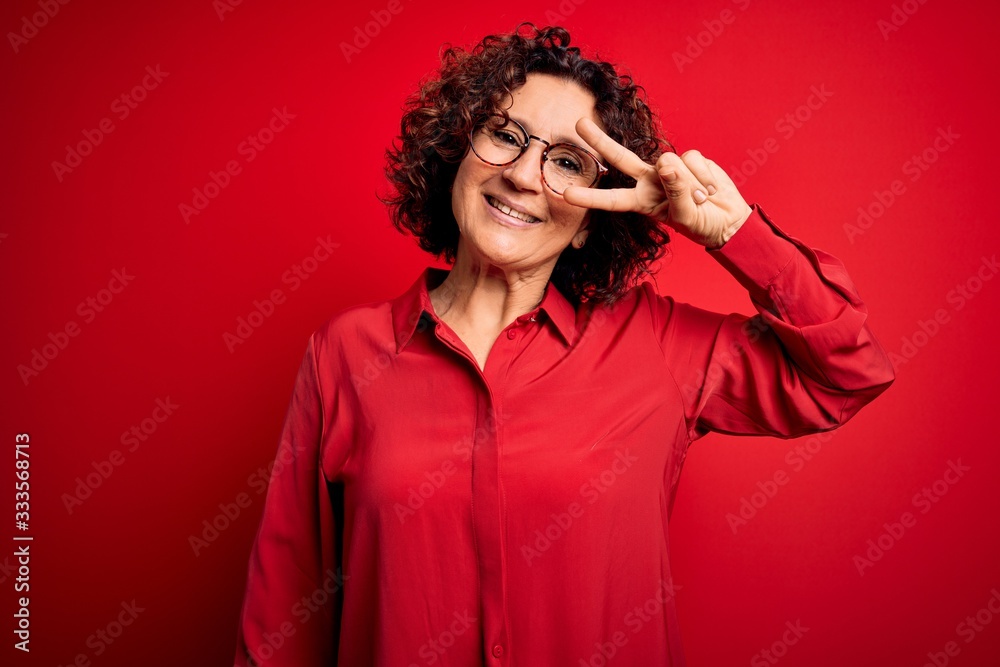 Middle age beautiful curly hair woman wearing casual shirt and glasses over red background Doing peace symbol with fingers over face, smiling cheerful showing victory