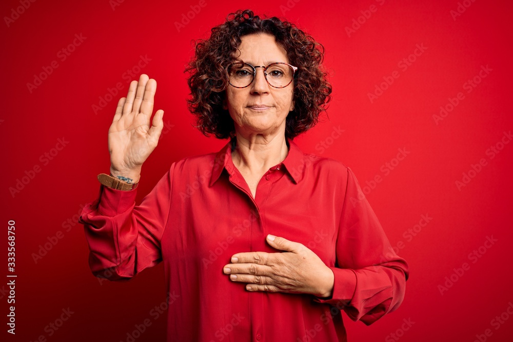 Middle age beautiful curly hair woman wearing casual shirt and glasses over red background Swearing with hand on chest and open palm, making a loyalty promise oath