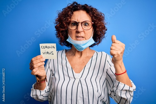 Middle age curly hair woman wearing medical mask holding reminder with corona virus message annoyed and frustrated shouting with anger  crazy and yelling with raised hand  anger concept