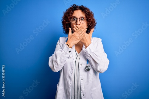 Middle age curly hair doctor woman wearing coat and stethoscope over blue background shocked covering mouth with hands for mistake. Secret concept.