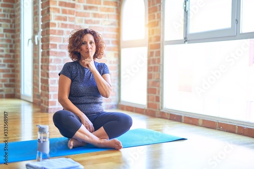 Middle age beautiful sportswoman wearing sportswear sitting on mat practicing yoga at home asking to be quiet with finger on lips. Silence and secret concept.