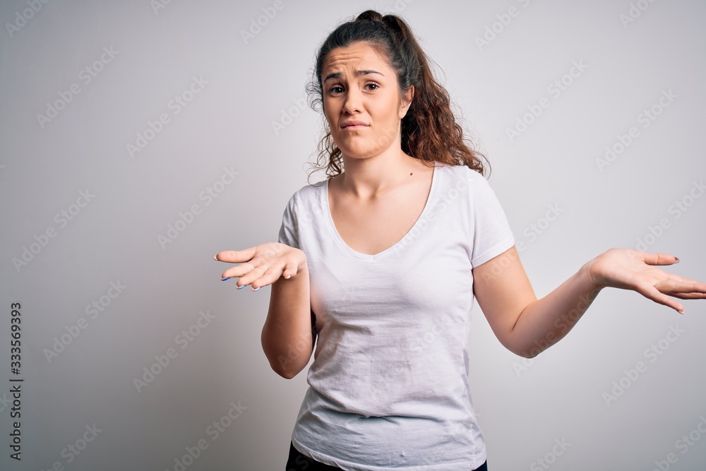 Young beautiful woman with curly hair wearing casual t-shirt standing over white background clueless and confused expression with arms and hands raised. Doubt concept.