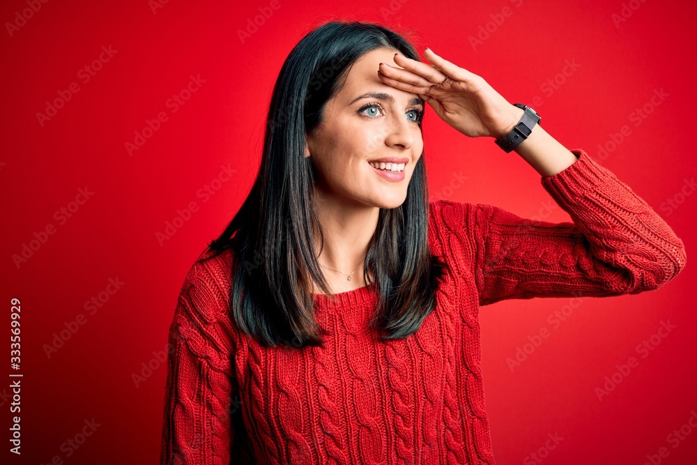 Young brunette woman with blue eyes wearing casual sweater over isolated red background very happy and smiling looking far away with hand over head. Searching concept. Young brunette woman with blue eyes wearing casual sweater over isolated red background very happy and smiling looking far away with hand over head. Searching concept.